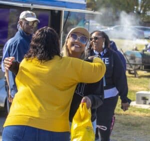 Two women hug warmly at an outdoor event, both smiling, while other attendees in the background laugh and converse. A food truck and barbecue grill with smoke rising are visible in the background.
