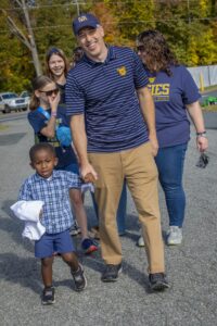 A smiling man wearing an N.C. A&T cap and polo holds hands with a young boy at an outdoor event. The boy is wearing a plaid shirt and holding a white cloth. In the background, other event attendees, including children, smile and walk together.