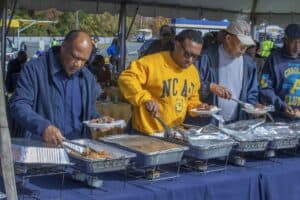 A group of men serving themselves food from a buffet table under a tent at an outdoor event. They are holding plates and using tongs to select food from chafing dishes. One man wears a bright yellow N.C. A&T sweatshirt, while others are dressed in casual attire.