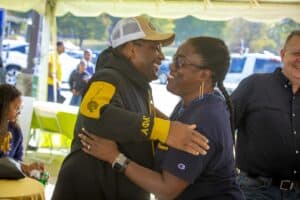A man and woman share a warm embrace under a tent at an outdoor event, both smiling. The man wears an N.C. A&T hat and hoodie with Alpha Phi Alpha branding, while the woman wears a navy event t-shirt. Other attendees are visible in the background, some seated and others standing.