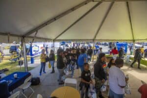 A wide view of an outdoor event under a large tent, showing attendees in line and engaging with various exhibits. People of all ages are gathered, holding event bags, with more activity visible outside, including booths and signage for the Agricultural Research Program.