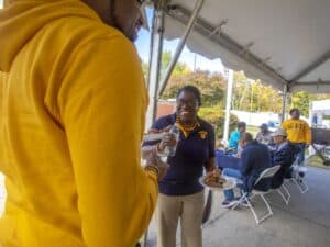 A woman wearing an N.C. A&T polo shirt smiles while holding a plate of food and a bottle of water under a tent at an outdoor event. In the background, attendees are seated at tables, enjoying their meals. Another person wearing a yellow hoodie stands in the foreground.