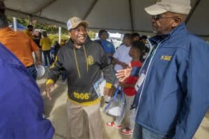A man wearing a black and gold hoodie with "1906" and an N.C. A&T cap smiles and reaches out to greet another man in a blue N.C. A&T jacket. They are under a tent at an outdoor event, with people, including children, gathered and interacting in the background.