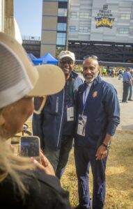 Two men wearing N.C. A&T jackets and event lanyards pose for a photo in front of Truist Stadium. A woman wearing a hat is seen in the foreground holding a phone to capture the picture. The stadium and event tents are visible in the background.