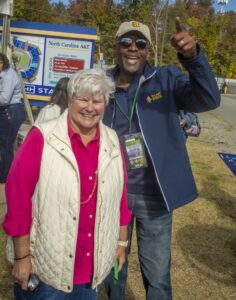 A man wearing an N.C. A&T cap and jacket raises his hand and smiles enthusiastically next to a woman in a white vest and pink shirt. They are standing outside near a stadium map sign for N