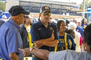 A group of people gathered under a tent at an outdoor event, including a man in an N.C. A&T cap and polo shirt standing with arms crossed, and a woman wearing an N.C. A&T jersey. Other attendees are visible in the background, with one person gesturing in the foreground.