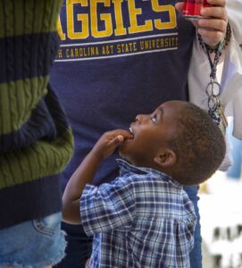 A young boy in a plaid shirt looks up and smiles while standing close to an adult wearing an N.C. A&T State University "Aggies" t-shirt. The adult holds a red soda can, and another person is partially visible on the left.