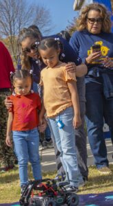 Two young girls stand close together, holding hands, while observing a small robotic vehicle on a colorful mat at an outdoor event. An adult leans in to speak to them, and another person wearing an N.C. A&T shirt stands nearby holding a phone.