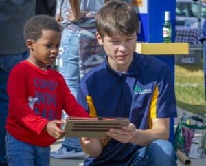 A young boy in a red shirt stands beside a young man in a blue and yellow polo shirt, both focused on a tablet held by the young man. They appear to be engaging in a demonstration or interactive activity at an outdoor event, with other attendees visible in the background.