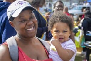 A smiling woman wearing a baseball cap holds a baby in her arms at an outdoor event. The baby, dressed in a light pink shirt, looks toward the camera with one finger in their mouth. People and event activities are visible in the blurred background.