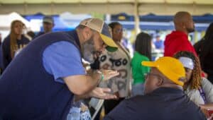 A man wearing a baseball cap leans forward, gesturing with his hands as he speaks to another man in a yellow cap seated at a table. Other attendees are visible in the background under a tent at an outdoor event.