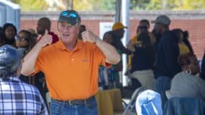 A man in an orange polo shirt and baseball cap gives two thumbs up under a tent. Several people are visible in the background, some seated and others standing.