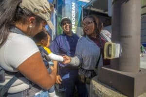 A woman holding a baby interacts with another woman outdoors near a booth. A man standing between them smiles while watching.