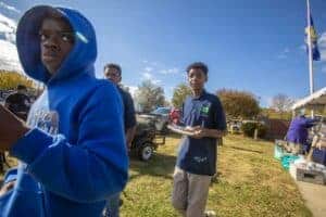 Two young men wearing blue shirts, one with a hoodie, walk outdoors during a sunny event. Another person is visible in the background holding a plate of food and a can of soda.