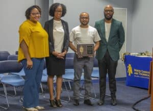 Four individuals, including Lauren Mayo, Ph.D., Devona Dixon, Ph.D., Arnab Bhowmik, Ph.D., and Kingsley Ekwemalor, Ph.D., pose for a group photo during an award ceremony. One individual holds a plaque. A table with the North Carolina A&T State University College of Agriculture and Environmental Sciences logo is visible in the background.