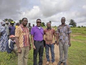 Four men stand together outdoors in a rural setting, smiling at the camera. They are surrounded by a group of people in the background, some wearing traditional attire. The sky is overcast, and the landscape features grassy fields and trees.