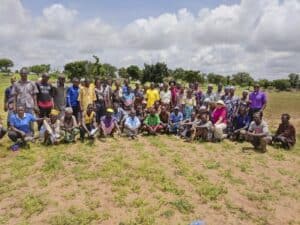 A large group of people, including farmers and community members, pose for a group photo in an open field under a partly cloudy sky. The group includes men and women of varying ages, with some standing and others crouching in front. Trees and greenery are visible in the background.