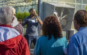 A speaker holds a microphone while addressing a group of attendees in front of a greenhouse. The speaker is wearing a gray polo shirt and gestures with one hand. The greenhouse equipment, including fans and hoses, is visible in the background. Attendees are seen from behind, listening attentively.