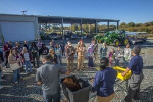 A group of farmers and attendees gathers outdoors at a farm during a field day event. Two presenters stand at a table displaying bins of soil and ginger. In the background, there are tractors, agricultural equipment, and a large open shelter. The event takes place under a clear blue sky.