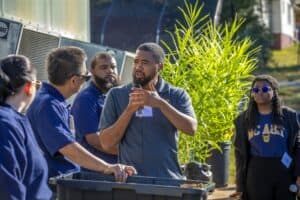 William Lashley, wearing a gray polo shirt, speaks into a microphone while gesturing with his hands during a presentation in front of a greenhouse. He is accompanied by his mentor, Guochen Yang, Ph.D., and other team members, who listen attentively. Potted plants, including ginger, are visible in the background, and another participant in NC A&T apparel observes the presentation.