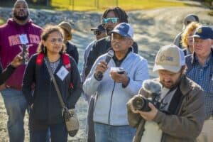 A man in a gray jacket holds a microphone and asks a question during a field day event. He is surrounded by other attendees, including a woman with a red backpack and a man holding a camera with a microphone attachment. The group stands on a gravel path under bright sunlight.