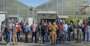 A large group of attendees gathers in front of greenhouses during a field day event. Guochen Yang, Ph.D., stands near the center, addressing the crowd and answering questions. Other presenters and participants are visible, with various agricultural displays set up in front of the greenhouse.
