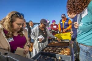 A group of attendees gathers around trays displaying ginger rhizomes and soil during a field day event. One participant examines the soil, while others take notes and observe closely. The event is held outdoors under a clear blue sky.
