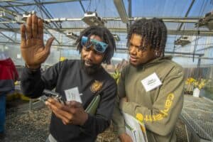 Farmers Darius McKoy and Avery Lucas discuss ginger farming inside a greenhouse during a field day event. Darius, wearing sunglasses and holding a smartphone, gestures as he talks, while Avery listens and holds event materials. The greenhouse structure and plant trays are visible in the background.