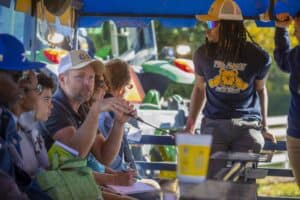 Michael Rayburn, wearing an NC A&T hat, speaks into a microphone while answering questions during the trolley transfer at a field day event. He is seated alongside other attendees, while a participant wearing an NC A&T Aggies shirt stands nearby. A tractor is visible in the background.