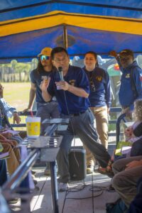 Guochen Yang, Ph.D., wearing an NC A&T polo shirt, speaks into a microphone while addressing attendees during a trolley ride at a field day event. He gestures with his hands as participants listen attentively. Other staff members in NC A&T attire stand nearby, and the background shows a tractor and farmland.