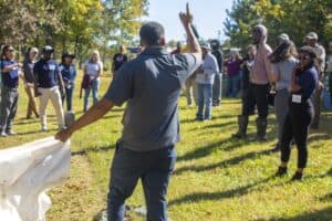 Ph.D. student William Lashley stands with his back to the camera, raising one hand as he explains the effects of colored shades in low tunnel ginger farming to a group of farmers. He holds a white tarp in his other hand. The group listens attentively in an open field surrounded by trees on a sunny day.