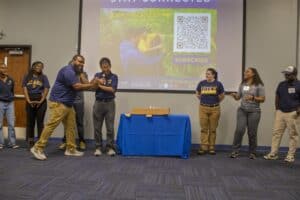 A group of people stands in a room during a presentation. One man is playfully handing a microphone to another, who smiles while holding it. Behind them, a projector screen displays a "Stay Connected" message with a QR code for subscribing to updates. A table with a blue tablecloth and a small box is in the center of the room. The participants wear casual attire, with some in North Carolina A&T shirts.