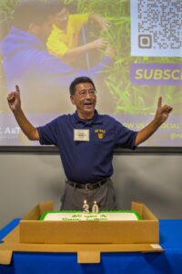 Guochen Yang, Ph.D., stands behind a cake with lit candles shaped as the number "30," celebrating his 30 years of research contributions. He raises both arms, smiling, while wearing a North Carolina A&T shirt. Behind him, a projector screen shows an image of people working with plants and a QR code for subscribing to updates.