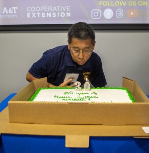 Guochen Yang, Ph.D., leans forward to blow out the "30" candles on a celebration cake, marking 30 years of tissue culture research. The cake is white with green trim and placed inside an open box. Behind him, a screen displays the North Carolina A&T Cooperative Extension logo and social media icons.