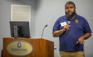 Extension specialist Trequan McGee, Ph.D., stands at a podium holding a microphone, speaking to attendees. He is wearing a navy blue North Carolina A&T Aggies polo shirt. The podium displays the North Carolina Agricultural and Technical State University emblem. A computer monitor and microphone are positioned on the podium.