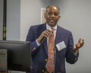 A man wearing a dark blue blazer, a paisley-patterned orange tie, and a checkered shirt speaks into a microphone while gesturing with his hand. He is standing near a Dell monitor in what appears to be a professional or conference setting. A name tag is visible on his chest.