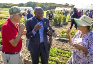 Three people engage in conversation outdoors during a small farms conference. The man on the left, wearing a red shirt and green cap, gestures while holding a small item. The man in the center, dressed in a suit and holding a camera, speaks animatedly. The woman on the right, wearing a patterned blouse and sun hat, responds while holding a sample of produce. In the background, people walk through garden beds filled with greenery and flowers, and greenhouses are visible in the distance.