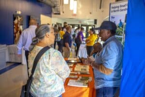A man in a denim shirt and cap speaks with a woman in a floral blouse at an orange-topped vendor table in a conference lobby. The table is covered with informational brochures and documents. In the background, other participants and vendors interact at additional tables, with some standing and others browsing materials. The space is bright with natural light, featuring a blue wall on the left and a banner on the right promoting support for small farmers in North Carolina. The atmosphere is lively and engaging.