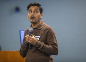 A man wearing a brown sweater and a name tag holds a microphone while speaking during a presentation. He appears engaged, with his gaze directed slightly upward. A blurred screen and desk are visible in the background, suggesting an indoor conference or lecture setting.