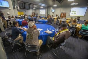 A large conference room with participants seated at round tables covered in blue and yellow tablecloths, attentively listening to a presenter at the front of the room. The presenter, standing near a podium, speaks in front of projected slides showing graphs and images related to livestock grazing. Some attendees take notes, while others look at their phones or conference materials. The room is brightly lit with ceiling lights, and additional screens display the presentation slides.