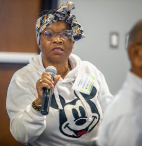 A woman wearing a white hoodie with a Mickey Mouse design and a patterned headscarf speaks into a microphone during a Q&A session. She is wearing glasses and a name tag that reads "Jamma Williams." The background is softly blurred, with another participant partially visible on the right.