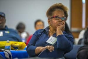 A woman wearing glasses with red and black frames and a dark blue shirt sits at a table, resting her chin on her hand, listening attentively during a conference session. She is wearing a name tag that reads "Margaret Harrison." A water bottle, a yellow bag, and papers are on the table in front of her. Other participants are visible in the softly blurred background.
