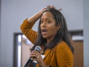 A woman with long black hair wearing an orange blazer holds a microphone and gestures with one hand raised above her head while speaking during a presentation. She is wearing a name tag, and her expression appears engaged and animated. The background shows a blurred doorway and wall, suggesting an indoor conference or workshop setting.