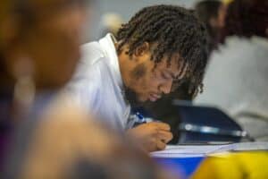 A young man with curly hair focuses intently while taking notes in a notebook during a conference session. He is holding a pen and appears engaged with the material. The background is softly blurred, showing other attendees and conference materials.