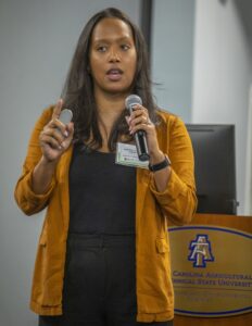 A woman with long black hair wearing an orange blazer speaks into a microphone while holding a presentation clicker in her other hand. She is wearing a name tag that identifies her as Andrea Gentry-Apple. In the background, a podium with the N.C. A&T State University logo is visible, along with a monitor, suggesting a professional conference or workshop setting.