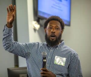 A man with a beard wearing a blue and white checkered shirt speaks into a microphone while raising one hand during a presentation. He is wearing a name tag that identifies him as Mark Paylor. A monitor displaying a presentation slide is visible in the background, suggesting a professional conference or workshop setting.