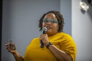 A woman wearing a yellow blouse and blue-framed glasses speaks into a microphone while holding a presentation clicker during a session. She has short, styled hair and is wearing a headband. The background is softly blurred, suggesting a professional conference or workshop setting.