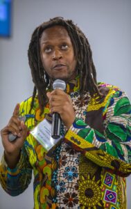 A man with long dreadlocks wearing a colorful patterned shirt speaks into a microphone while holding a presentation clicker in his other hand. He is wearing a name tag that identifies him as Michael Carter Jr. The background is softly blurred, suggesting an indoor conference or workshop setting.