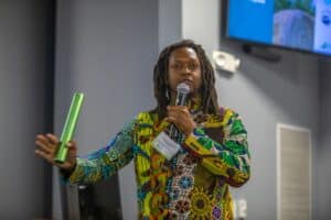 A man with long dreadlocks wearing a colorful patterned shirt speaks into a microphone while holding a green baton in his other hand. He is wearing a name tag identifying him as Michael Carter Jr. A monitor displaying a presentation slide is visible in the background, indicating a professional conference or workshop setting.