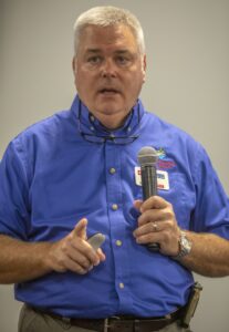 A man wearing a blue button-up shirt with a North Carolina State University Cooperative Extension logo speaks into a microphone during a presentation. He holds a presentation clicker in his other hand and has glasses resting on his shirt collar. His expression is focused as he gestures slightly with his hand. The background is neutral, suggesting an indoor conference setting.